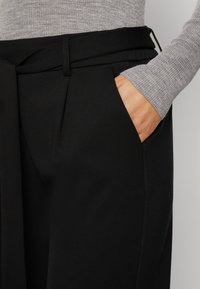 Black trousers with a relaxed fit, featuring a belted waist, side pocket, and a smooth texture, contrasted by a gray ribbed top.