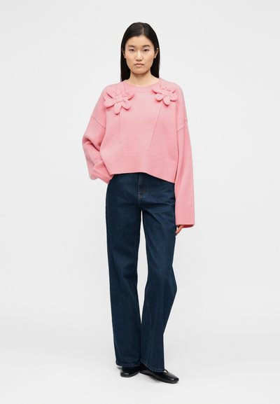 Ροζ πλεκτό πουλόβερ με άνετη, φαρδιά εφαρμογή, διακοσμημένο με τρισδιάστατα λουλούδια στους ώμους και ριμπ τελείωμα. Συνδυάζεται με σκούρο τζιν.
