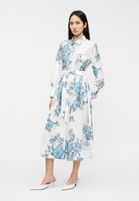 Λευκό φόρεμα με λουλούδια που διαθέτει μπλε τριαντάφυλλα, μακριά μανίκια, κουμπιά μπροστά και ζώνη από ύφασμα. Συνδυασμένο με λευκά μυτερά παπούτσια χωρίς κορδόνια.