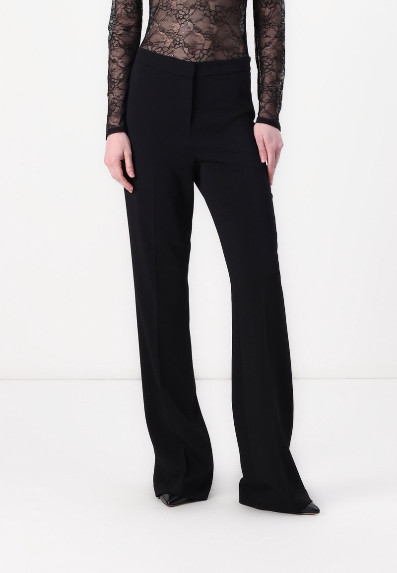Pantalon large taille haute noir porté avec des chaussures noires à bouts pointus, assorti d'un haut en dentelle transparente à manches longues.