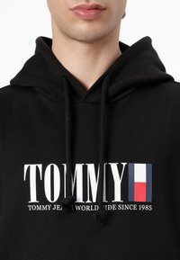 Crna dukserica s bijelim "TOMMY" logotipom, plavim i crvenim pravokutnim detaljem, vezicom oko vrata, mekim materijalom i opuštenim krojem.