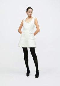 Λευκό φόρεμα με υφή, στενή γραμμή στο μπούστο και φαρδιά φούστα, με διακοσμητικές τσέπες, φορεμένο με μαύρες κάλτσες και μυτερές γόβες.