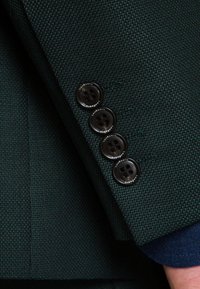 Mrok zielona rękaw marynarki z teksturowanym materiałem, cztery czarne guziki o okrągłym wzorze i niebieski mankiet wystający  spod spodu.