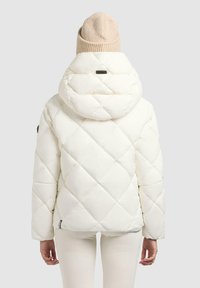 Witte gewatteerde pufferjack met een grote kap, voorzien van een diamantpatroon en een klein zwart logo nabij de zoom. Lichtbeige gebreide beanie bovenop.
