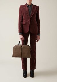Person wearing a burgundy corduroy suit and black boots holding a large brown textured handbag with black handles and gold clasp.