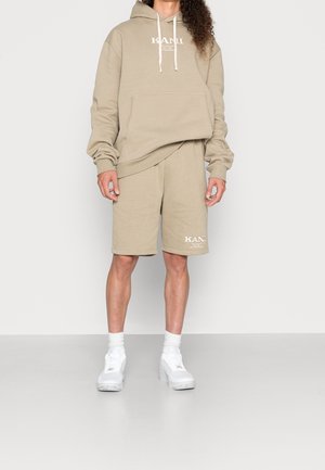 Beiger Hoodie mit Fronttasche und Logo, kombiniert mit passenden Shorts. Getragen mit weißen Sneakers und Socken. Lässiger, entspannter Schnitt.