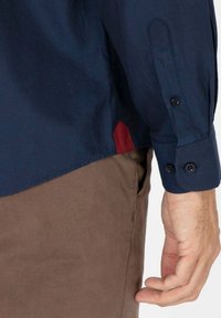Chemise à manches longues bleu marine avec des poignets boutonnés, comportant un accent rouge sur l'ourlet latéral et un tissu lisse et texturé.