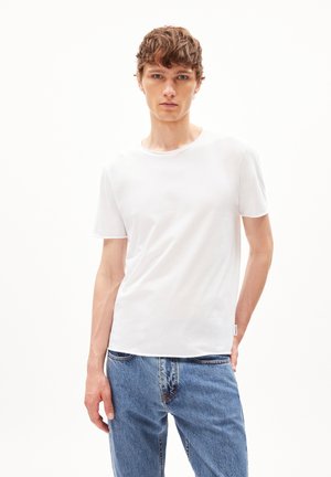 Jonge man draagt een eenvoudig wit T-shirt met korte mouwen en blauwe spijkerbroek, staat tegen een witte achtergrond en kijkt recht vooruit.