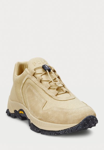 Beige velúrbőr és hálós sneaker, textúrált felülettel, fekete gumitalppal és elasztikus fűzőkkel. A nyelvén logó található, és támogató kivitel jellemzi.