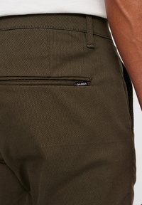 Kaki hlače s teksturiranim materijalom, imaju stražnji džep s crnom GABBA oznakom. Kroj je prilagođen s čistim dizajnom.