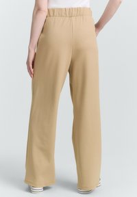 Pantaloni beige a gamba larga con cintura elastica, texture liscia e tasche laterali, abbinati a sneakers bianche basse.