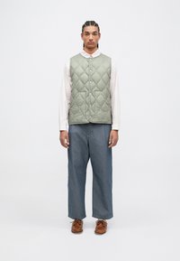 Rūpīgi izšūts, gaiši zaļš veste ar pogu aizdari, valkāta pār rozā krekli. Kombinēta ar brīviem ziliem džinsu biksēm un brūnām kurpēm.