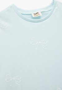 Camiseta de manga corta azul claro con lazo blanco bordado. Textura de algodón suave con cuello redondo y etiqueta en el interior.