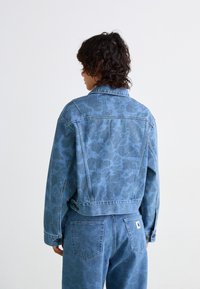 Veste en denim bleu clair avec une texture à motif subtil. Dotée d'un col classique et de fermetures à boutons. Associe à un jean assorti.