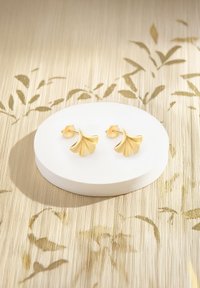 Gli orecchini a perno a forma di foglia di ginkgo in oro presentano un design testurizzato con una finitura lucida, posizionati su una base circolare bianca liscia contro uno sfondo decorato.
