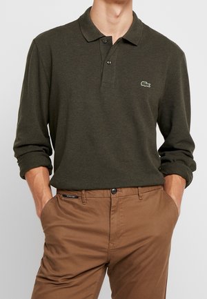 Mand iført mørkegrøn langærmet poloshirt med krokodillelogo og brune bukser, hænderne i lommerne, foran ensfarvet baggrund.