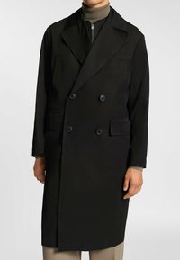 Czarny dwurzędowy płaszcz trencz z dużymi klapami, teksturowana tkanina, dwie przednie kieszenie i czarne guziki, noszony na ciemnej golfie.