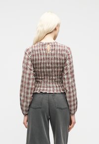 Γυναίκα με μακριά ξανθά μαλλιά, φορώντας κόκκινη και λευκή πουα μπλούζα με σφηκοφωλιά και γκρι φαρδιά τζιν ψηλόμεσα, εμφανίζεται από την πίσω πλευρά.