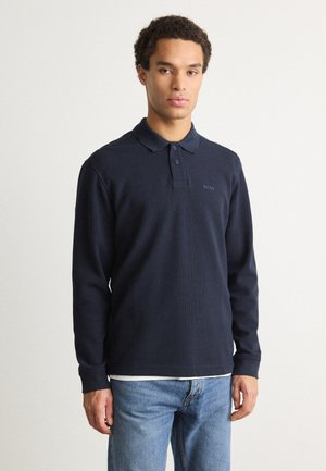 Polo cu mânecă lungă de culoare navy, cu guler și plastron cu trei nasturi, textură ribbed și subtile logo "BOSS" pe piept, asortat cu blugi deschiși.