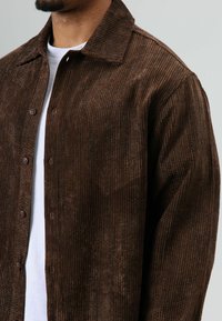 Bruine corduroy blouse met knopen, verticale geribbelde textuur, spitse kraag en bruine knopen. Gedragen over een wit T-shirt.