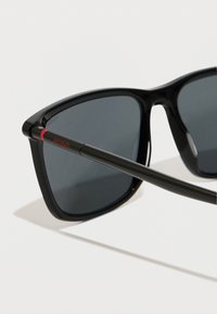 Mustad mustad prillid tumedate läätsa, läikivast plastist raam ja peen must vars, millel on väike punane 'HUGO' logo detail.