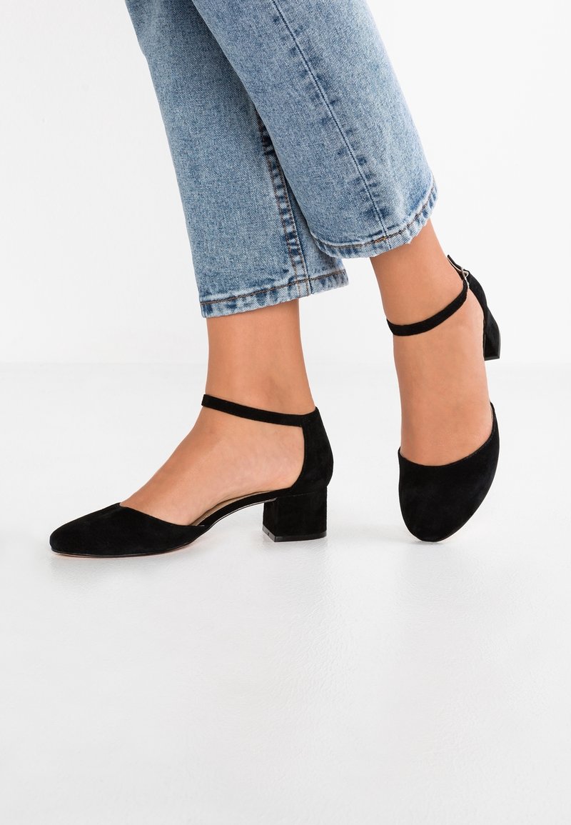 Femme portant des chaussures à talons carrés en daim noir avec des brides autour de la cheville, associées à un jean en denim bleu retroussé, debout sur un sol blanc.