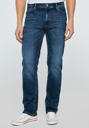 Man wearing dark blue straight-leg jeans and white low-top sneakers standing against plain background.