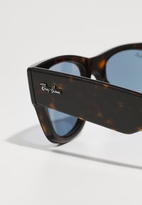 Ochelari de soare tortoiseshell maro cu lentile albastre. Au o ramă pătrată mare și un logo argintiu Ray-Ban pe sprijin. Textură netedă.