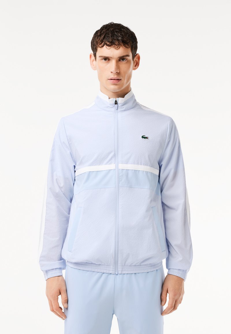 Veste zippée bleu clair avec un col montant, bandes blanches sur la poitrine et tissu texturé. Présente un petit logo vert sur le côté gauche.