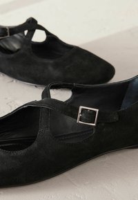 Zwarte suède schoenen met een puntige neus en een kruisriemontwerp. Kenmerken zijn een kleine zilveren vierkante gesp en een gladde binnenvoering.