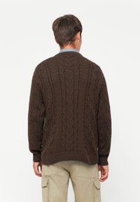 Uomo di spalle che indossa un maglione a trecce marrone scuro e pantaloni cargo beige su uno sfondo chiaro e uniforme.