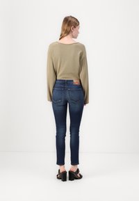 Suéter de oliva de manga larga, jeans de mezclilla azul ajustados con un lavado oscuro y sandalias negras de tacón grueso. Etiqueta trasera visible en los jeans.