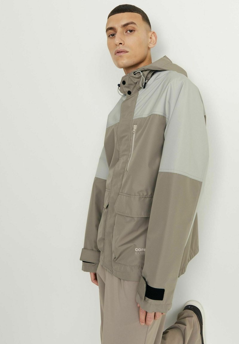 Lichte, waterafstotende jas in beige en grijs. Voorzien van een ritsborstzak, verstelbare capuchon en contrasterende zwarte manchetten.