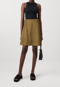 Top negro sin mangas, falda acampanada de pana verde olivo y zapatos negros de slip-on. Modelo sosteniendo un pequeño bolso negro.