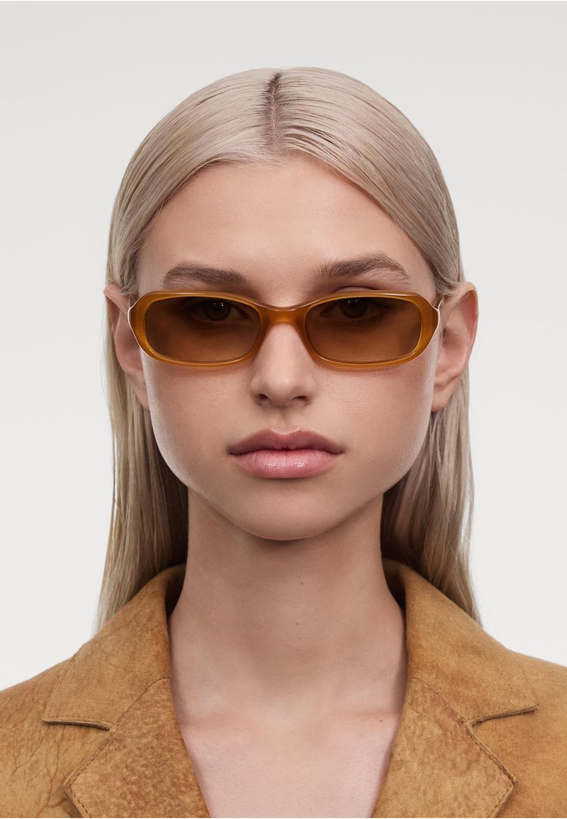 Schlanke braune Sonnenbrille mit rechteckigem Rahmen, transparenten Bügeln und weichen, glänzenden Gläsern. Getragen von einer Person mit glattem, blonden Haar.