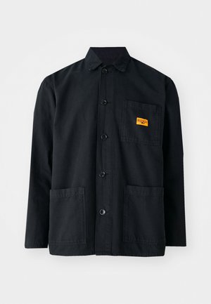 Veste de travail en coton noir avec un devant à boutons, deux poches avant et une petite étiquette jaune sur la poche poitrine. Design simple et utilitaire.
