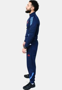 Chándal atlético navy con rayas azul claro, acentos del logo en rojo, diseño ajustado, combinado con zapatillas negras, mostrando el perfil lateral.