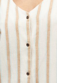 Λευκό ύφασμα με κάθετες μπεζ και ανοιχτού γκριζοπράσινου γραμμές, με V-λαιμόκοψη και τρία σκουροκάστανα κουμπιά στο μπροστινό μέρος.