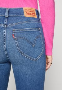 Jeans de mezclilla azul con una cintura alta, que presentan bolsillos traseros prominentes con un diseño de costura en forma de corazón. Parche con el logo de Levi's en la cintura.