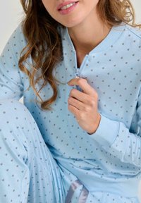 Mujer con cabello rizado que lleva un pijama de lunares azul claro, sosteniendo la cremallera de la parte superior cerca de su pecho.