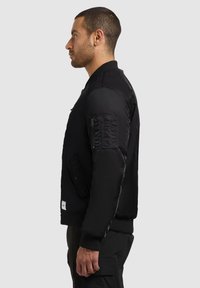 Giubbotto bomber nero con finitura satinata, polsini e colletto a coste, tasche laterali e tasca con zip sulla manica. Presenta una silhouette aderente.