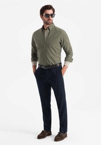 Olijfgroene shirt, marineblauwe geruite broek, bruine schoenen en zonnebril. Het shirt heeft een zak; de broek heeft een getailleerde pasvorm.