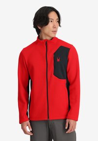 Giacca rossa texturizzata con accenti neri, chiusura con zip e tasca sul petto sinistro con logo a ragno. Maniche a lunghezza intera.