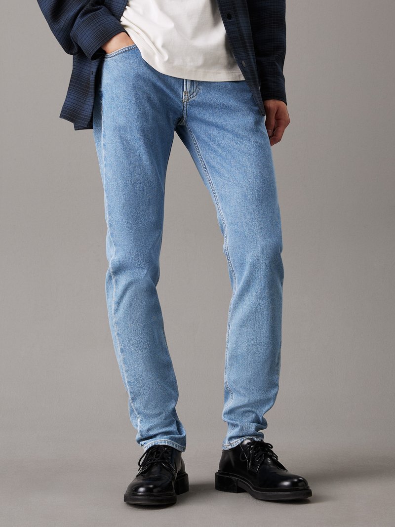 Vaqueros azul claro de mezclilla con corte ajustado. Presentan un diseño clásico de cinco bolsillos y costuras detalladas. Combinados con zapatos negros.