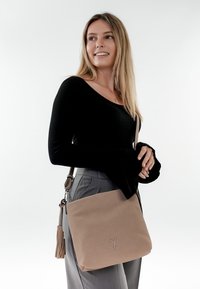 Frau mit langen blonden Haaren, trägt ein schwarzes Oberteil und graue Hosen, eine beige Umhängetasche mit Quasten-Detail, lächelt und schaut zur Seite.