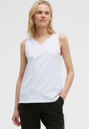 Blonde Frau mit ärmellosem weißen Oberteil und schwarzen Hosen, steht mit einer Hand in der Tasche vor einem schlichten Hintergrund.