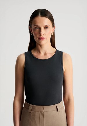 Kobieta o prostych brązowych włosach, ubrana w czarny bezrękawnik i beżowe spodnie z wysokim stanem, stojąca na jednolitym jasnoszarym tle.