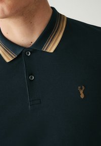 Mörkgrön polo skjorta med en krage i guldrand, två svarta knappar och en liten guldhjort-logga på bröstet. Texturerat tyg.