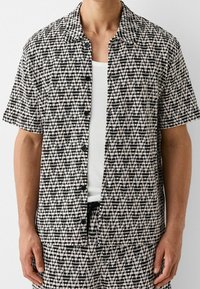 Hombre vestido con una camisa de botones de manga corta de patrón en blanco y negro sobre una camiseta de tirantes blanca con textura acanalada, y pantalones cortos a juego con el mismo patrón.