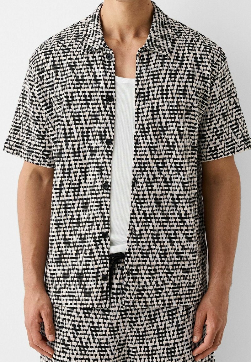 Hombre vestido con una camisa de botones de manga corta de patrón en blanco y negro sobre una camiseta de tirantes blanca con textura acanalada, y pantalones cortos a juego con el mismo patrón.